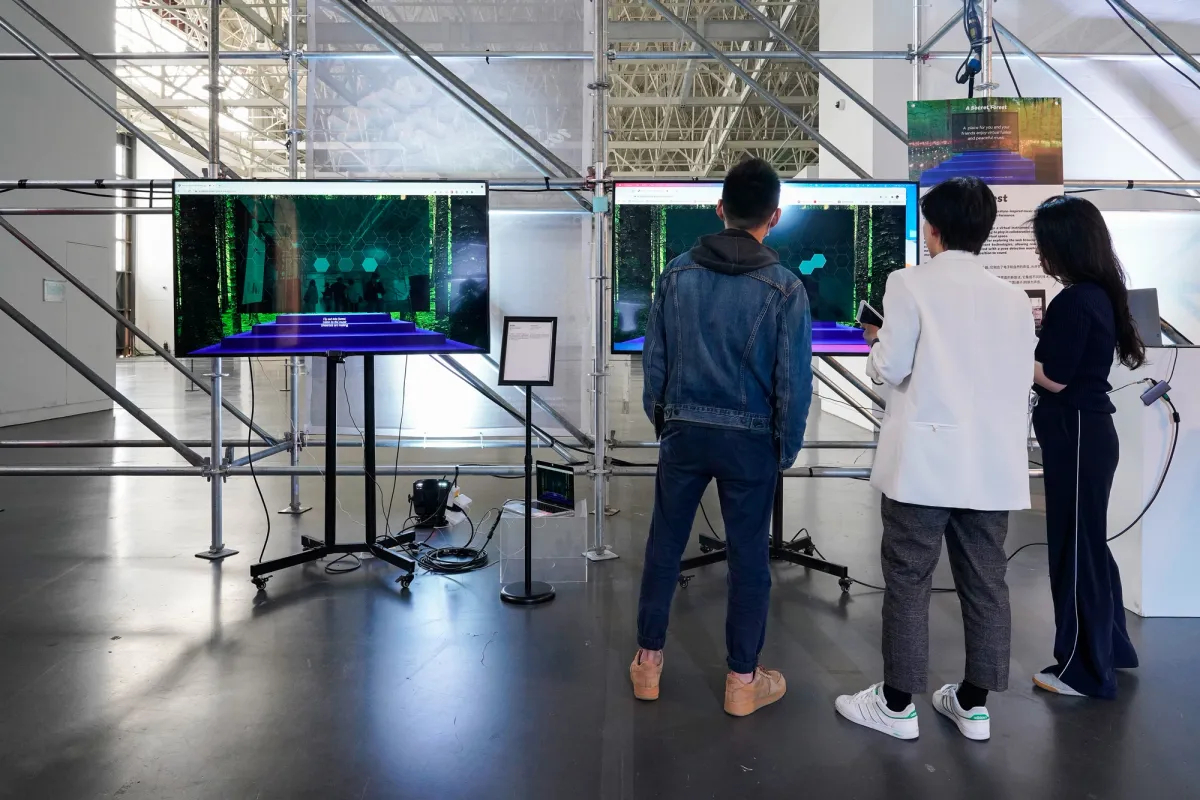Two digital monitors displaying art installations in a spacious museum gallery. Three individuals stand observing the screens.