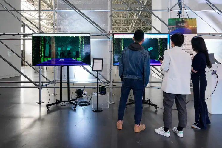 Two digital monitors displaying art installations in a spacious museum gallery. Three individuals stand observing the scre...