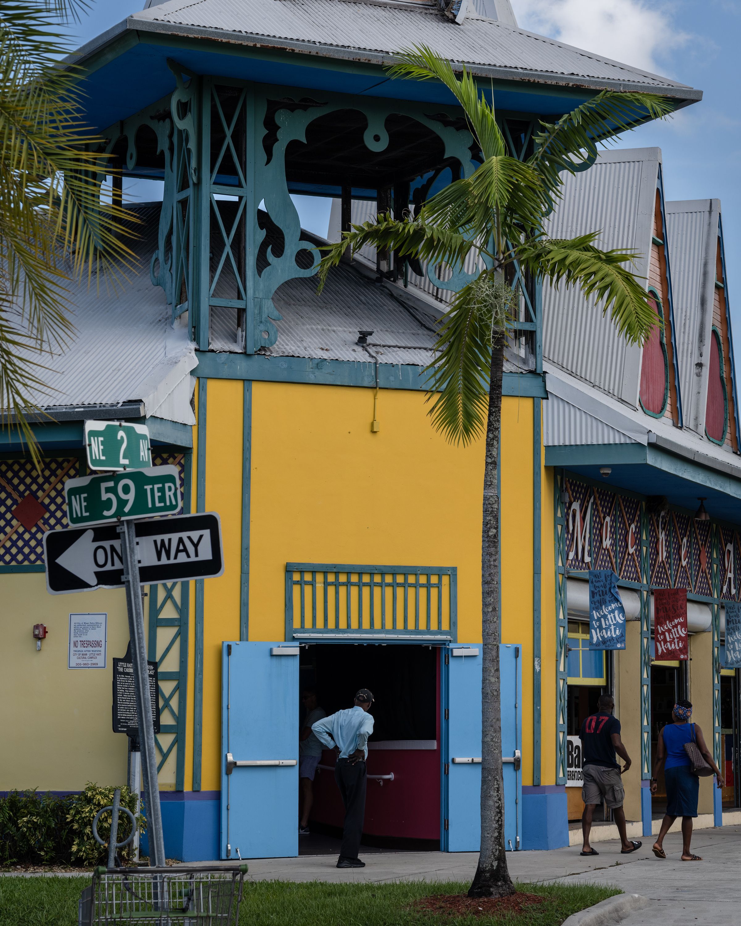 A vibrant yellow and blue building at the Little Haiti Cultural Center in Miami, with palm trees surrounding it on a clear, sunny day. A street sign and a one-way sign are positioned in the front.