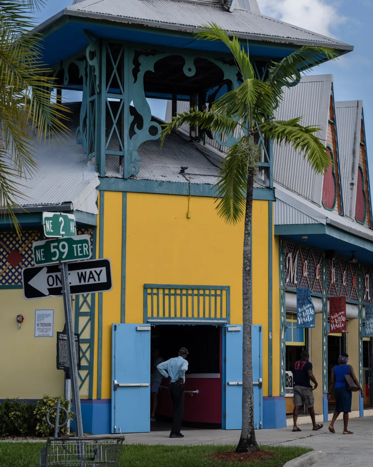 A vibrant yellow and blue building at the Little Haiti Cultural Center in Miami, with palm trees surrounding it on a clear, sunny day. A street sign and a one-way sign are positioned in the front.