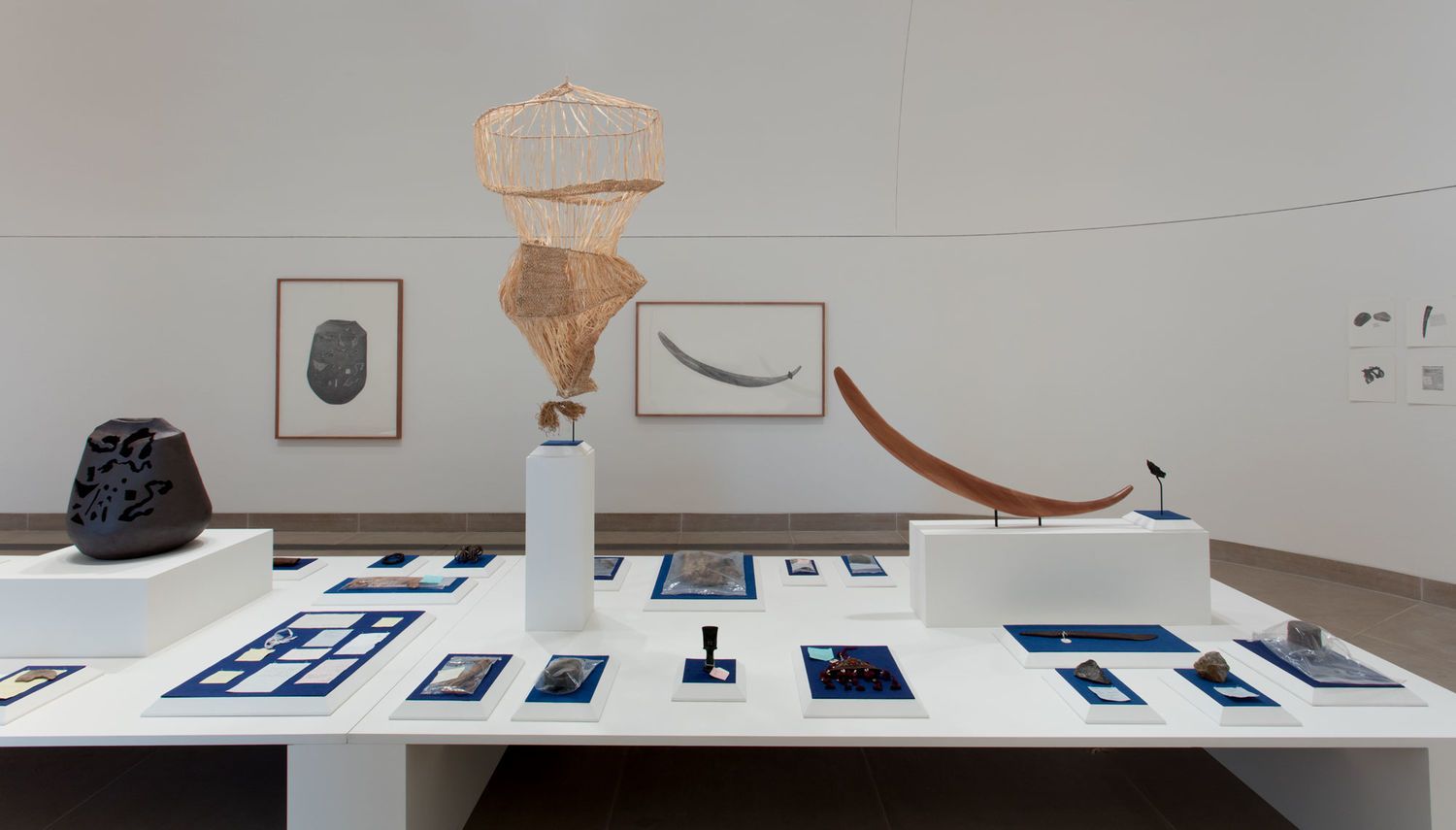 Exhibition in a gallery featuring rocks, a vase, and framed artworks hanging on the wall and laid out on a table.