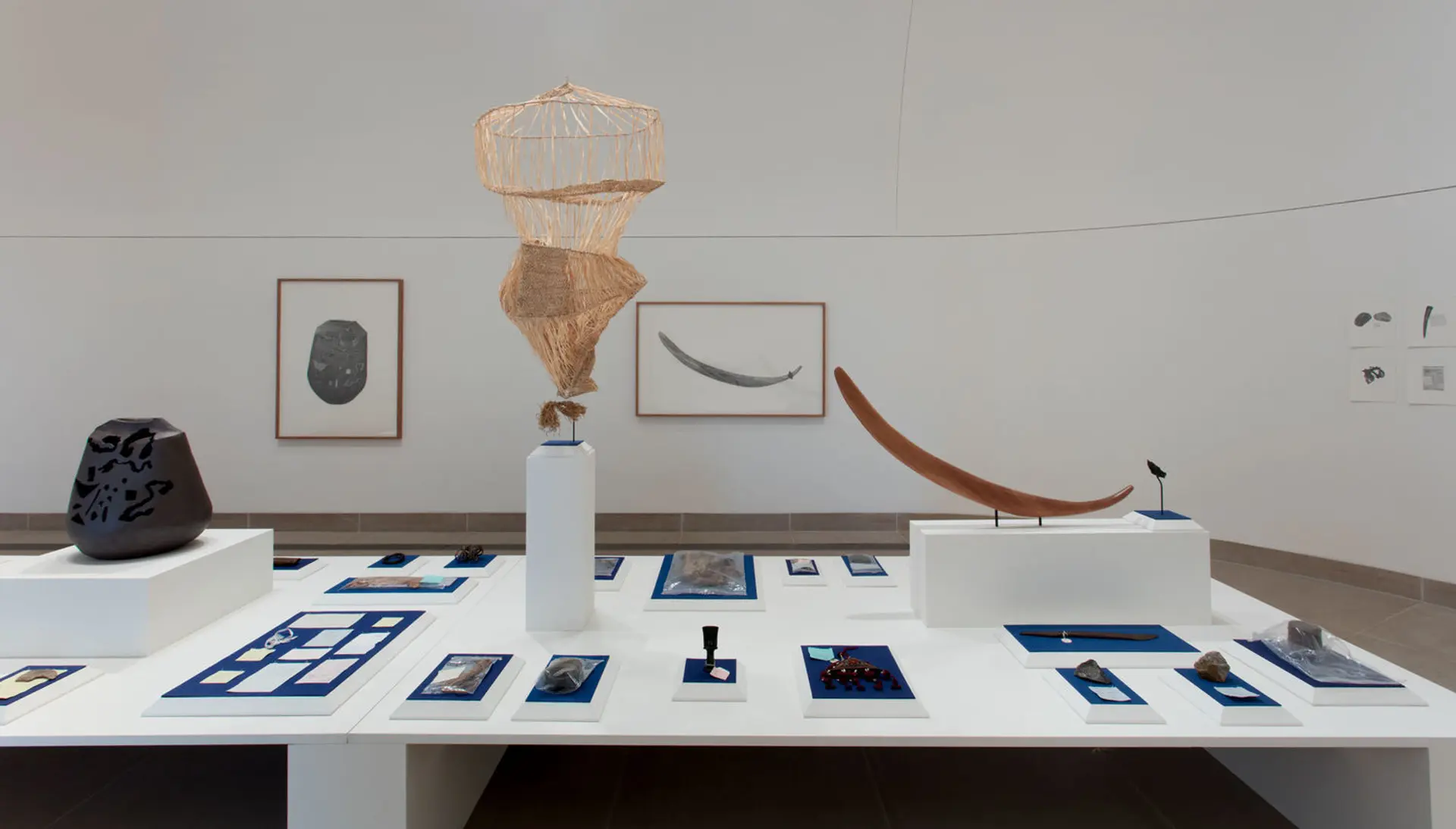 Exhibition in a gallery featuring rocks, a vase, and framed artworks hanging on the wall and laid out on a table.