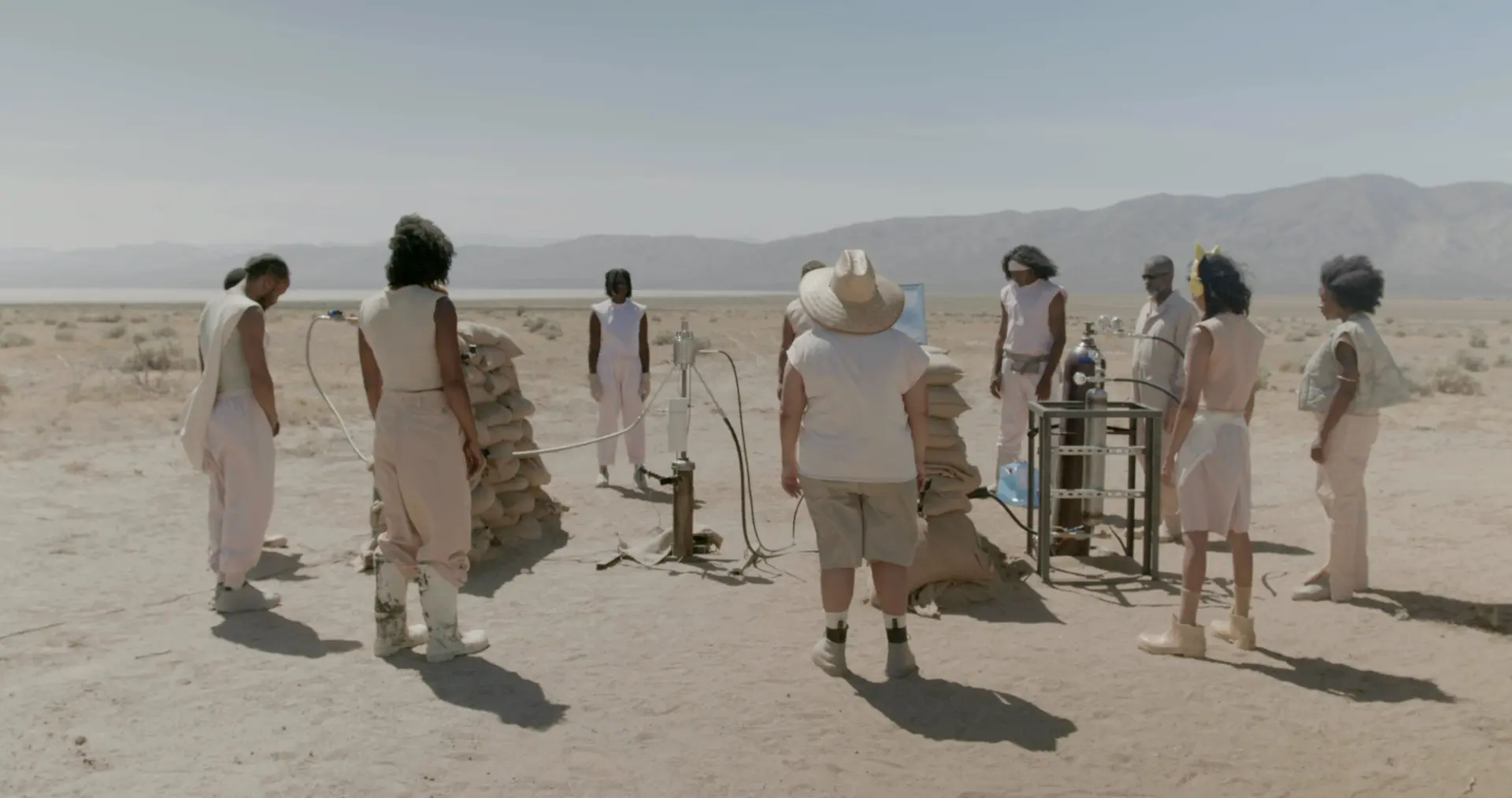A group of people stands in a desert circle around equipment and sandbags, engaging in a ritual or gathering.