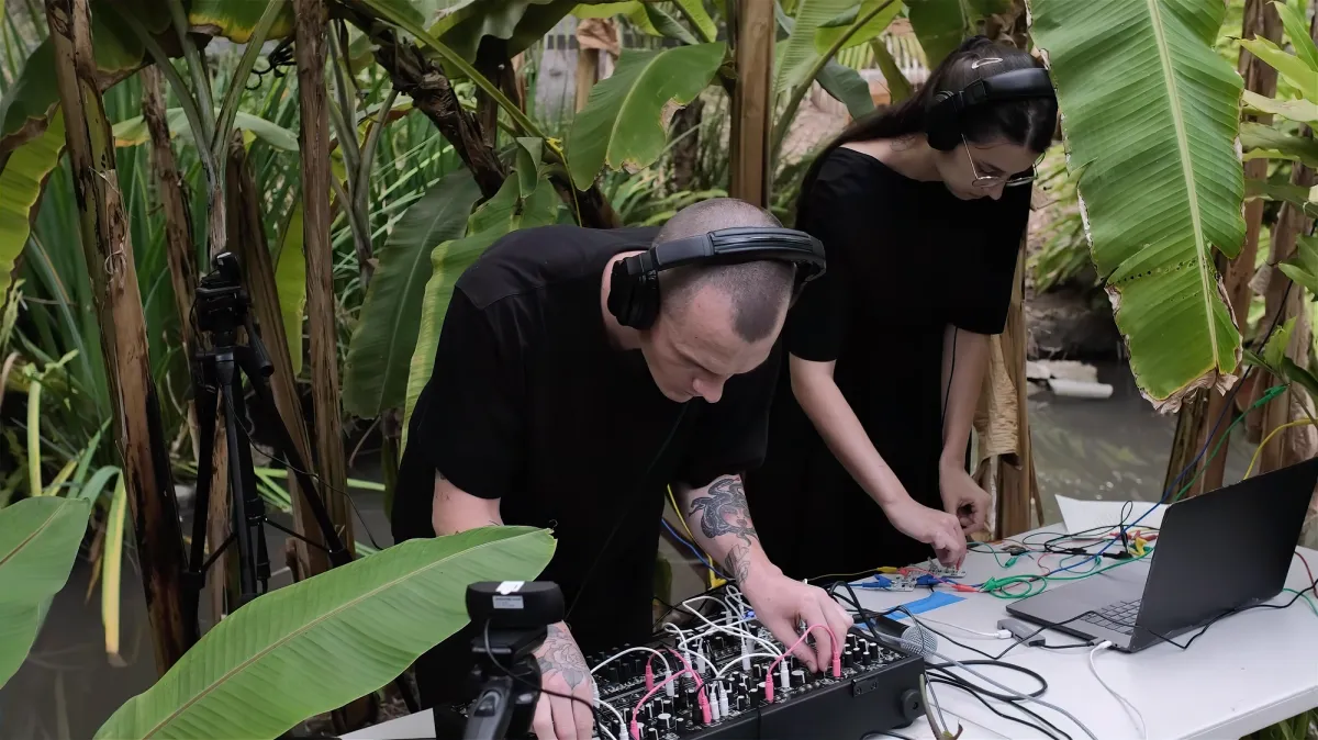 Ivana Dama and Clinton Van Arnam are working together on audio electronics and a laptop, amidst a lush environment of large green plants. They are connecting these plants to a midi controller to trigger a morse code message.