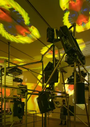 Projection equipment and pipes fill a room, casting vibrant yellow, blue, and red projections onto the ceiling and wall.