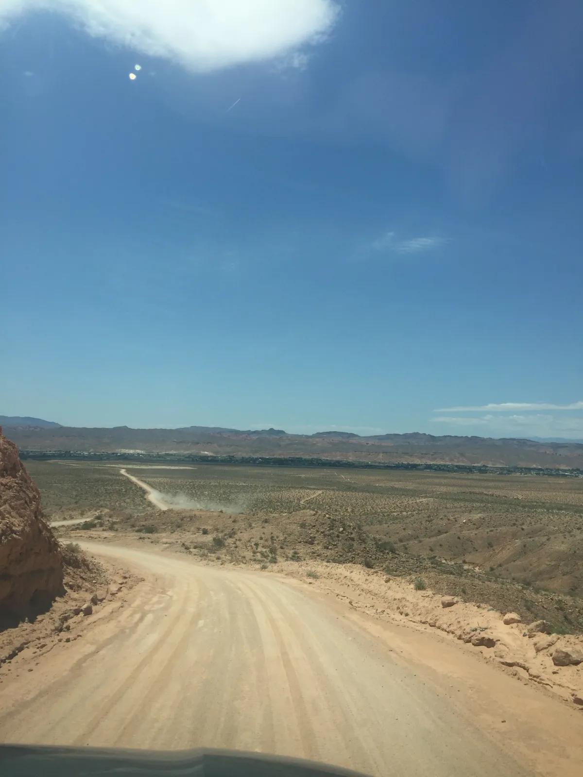 A sunlit road cuts across a desert landscape facing a clear horizon under a sky scattered with clouds.