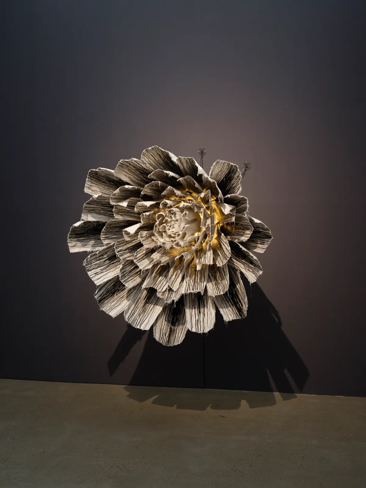 A wall-mounted art installation, reminiscent of a blooming flower, with a bright light at the center against a black wall.
