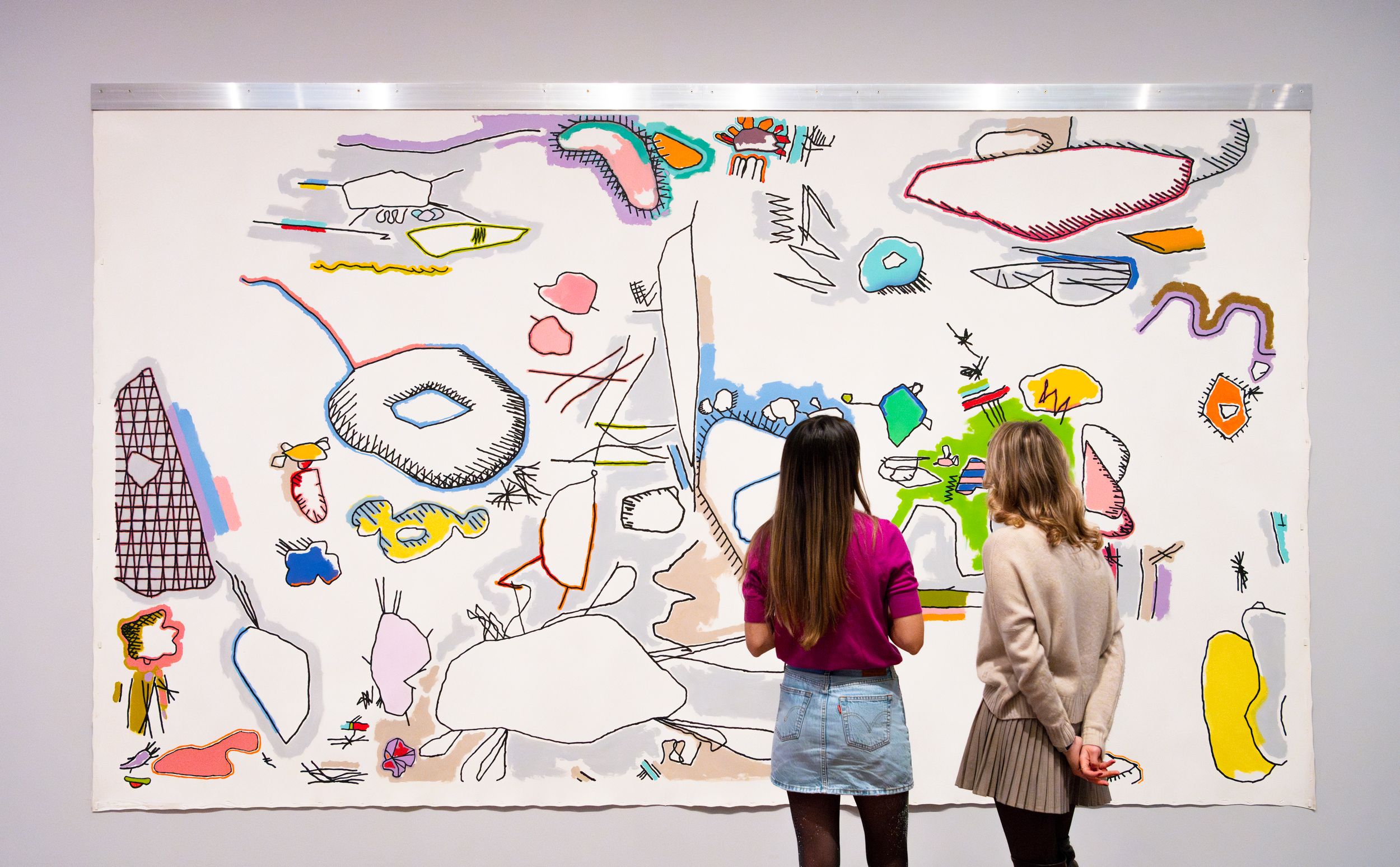 Two people stand closely in front of a large wall-mounted artwork filled with colorful, abstract doodle-like forms outline...
