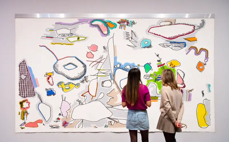 Two people stand closely in front of a large wall-mounted artwork filled with colorful, abstract doodle-like forms outline...