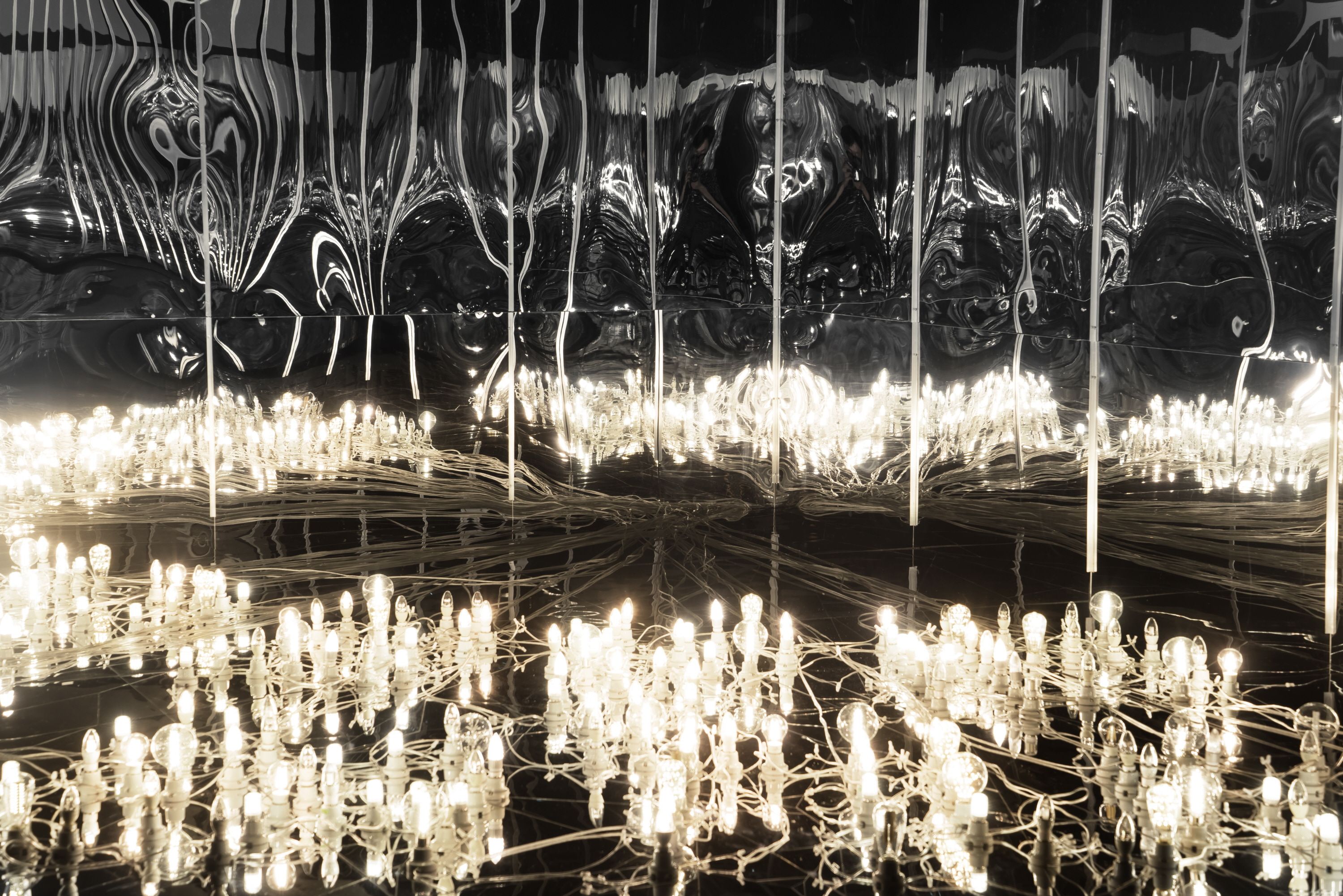 An array of wired yellow lights arranged on the floor of a room, casting luminous reflections onto the surrounding mirror ...