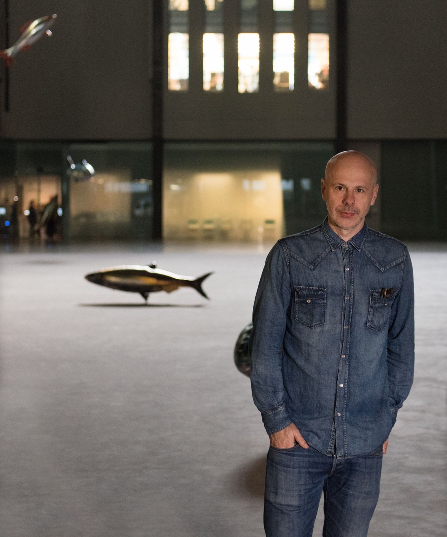 A person in jeans and a denim shirt stands in front of an art installation featuring a variety of fish.