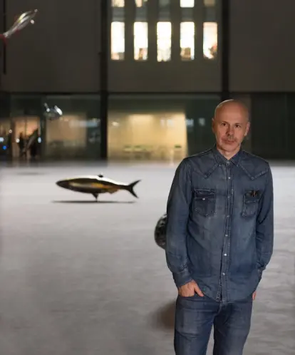 A person in jeans and a denim shirt stands in front of an art installation featuring a variety of fish.