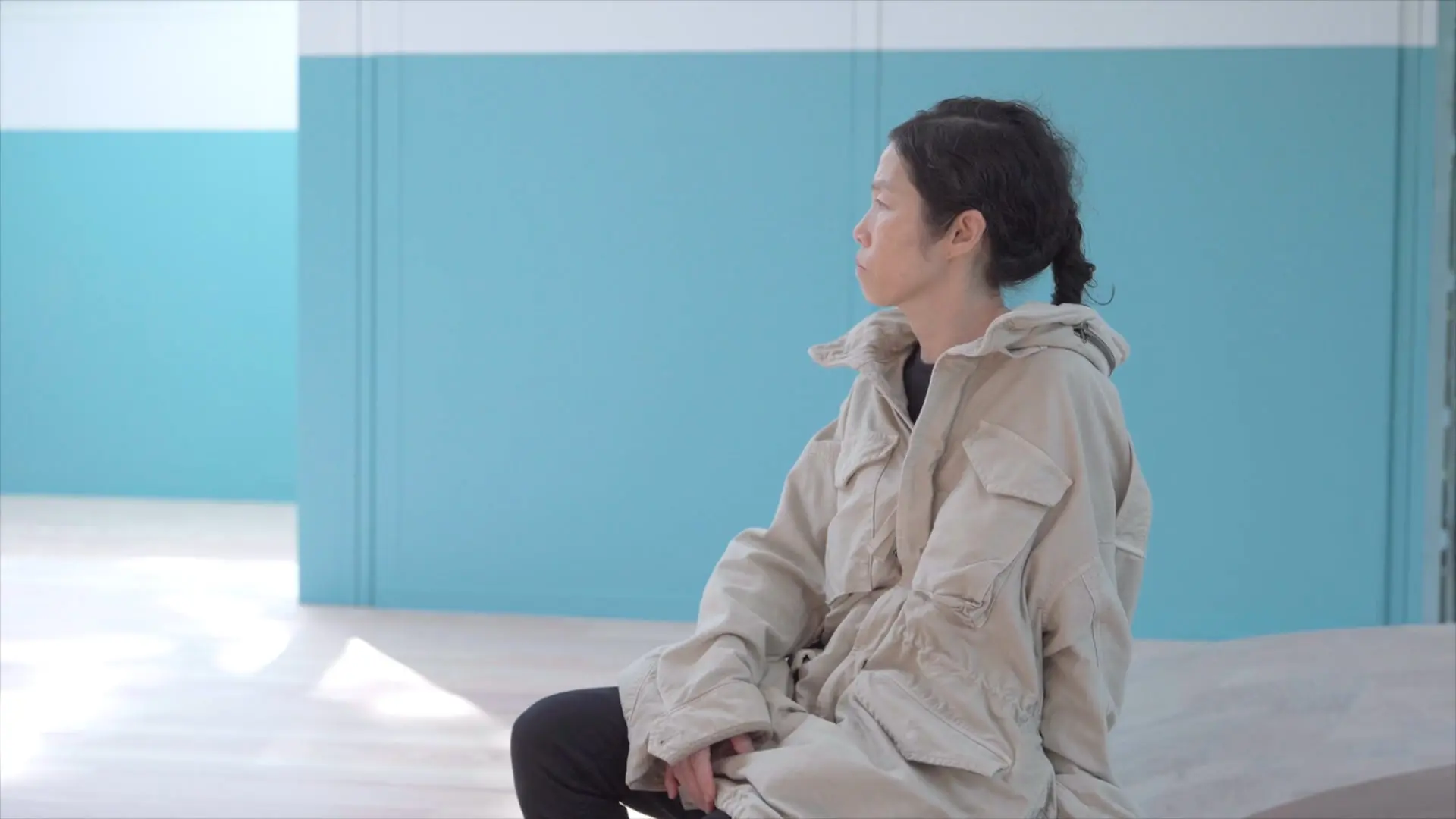 A woman in a beige jacket sits pensively in a room with light blue walls.