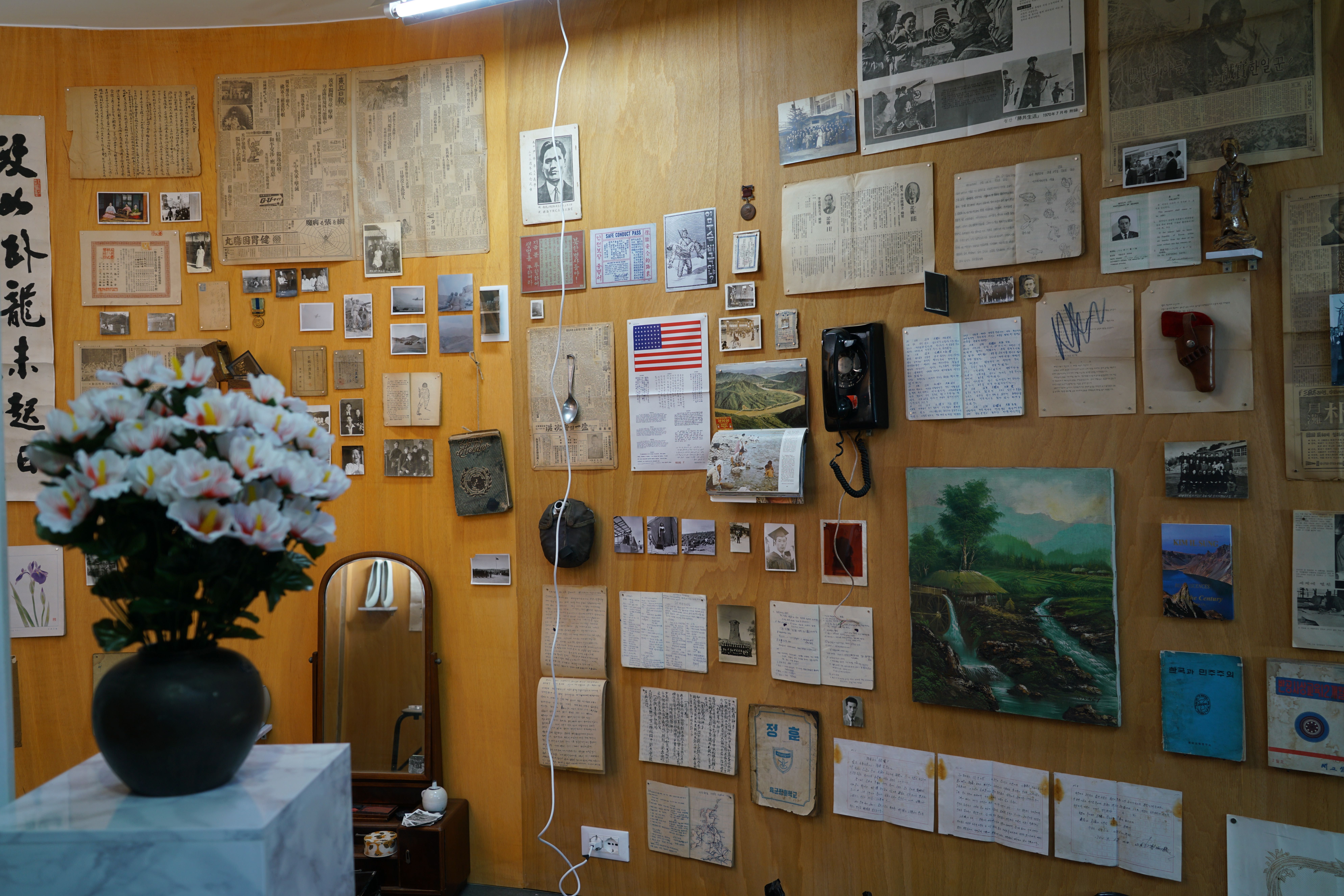 A wooden wall cluttered with an eclectic mix of newspapers, monochrome photos, handwritten notes, and painted images. An a...