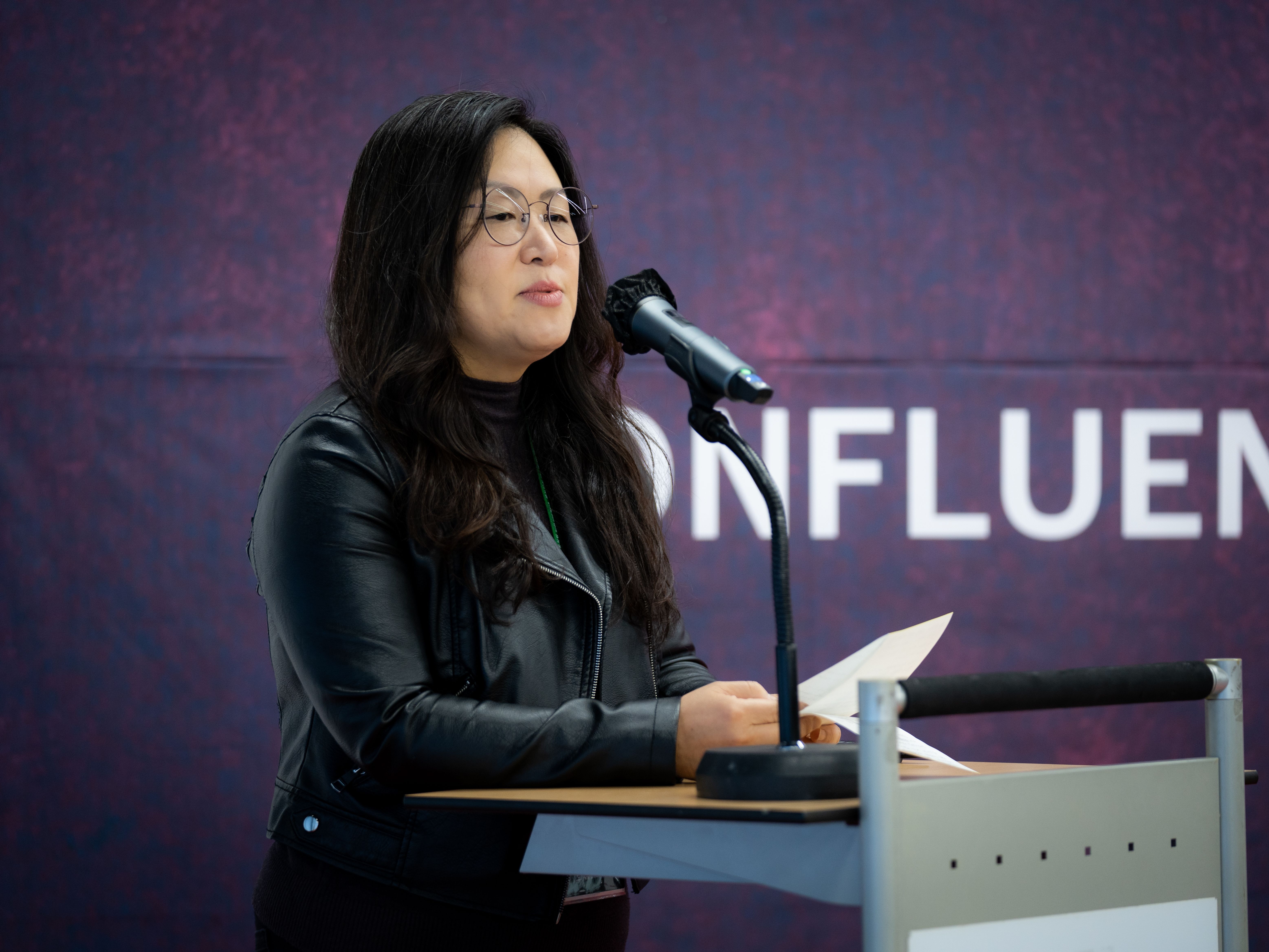 A person in a black leather jacket, addresses an audience behind a microphone stand. behind a microphone stand.