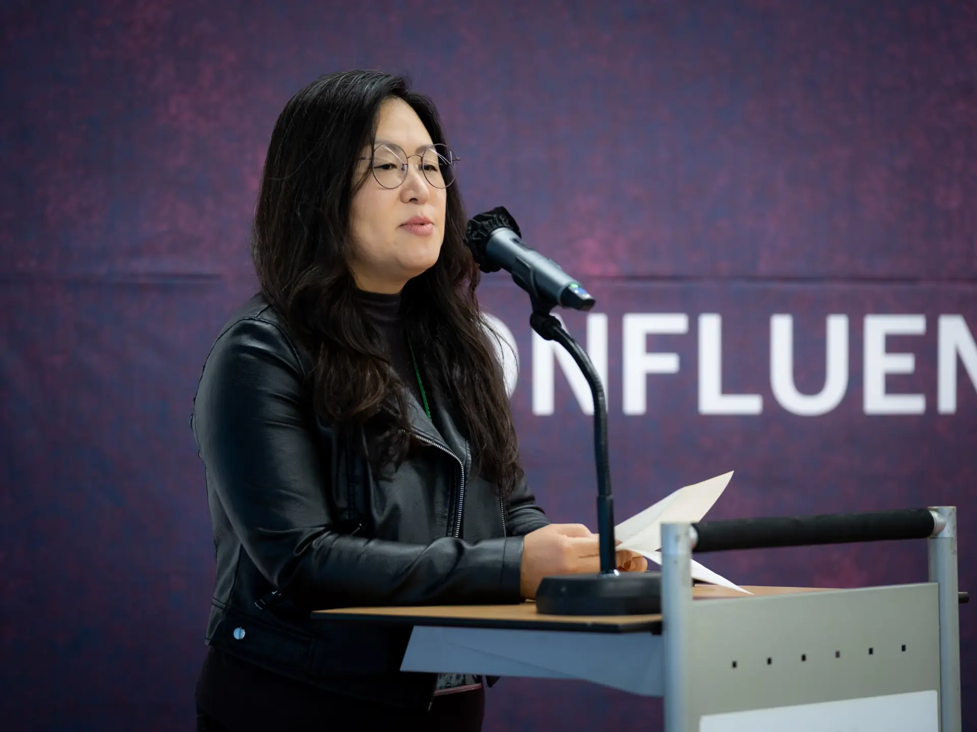 A person in a black leather jacket, addresses an audience behind a microphone stand. behind a microphone stand.