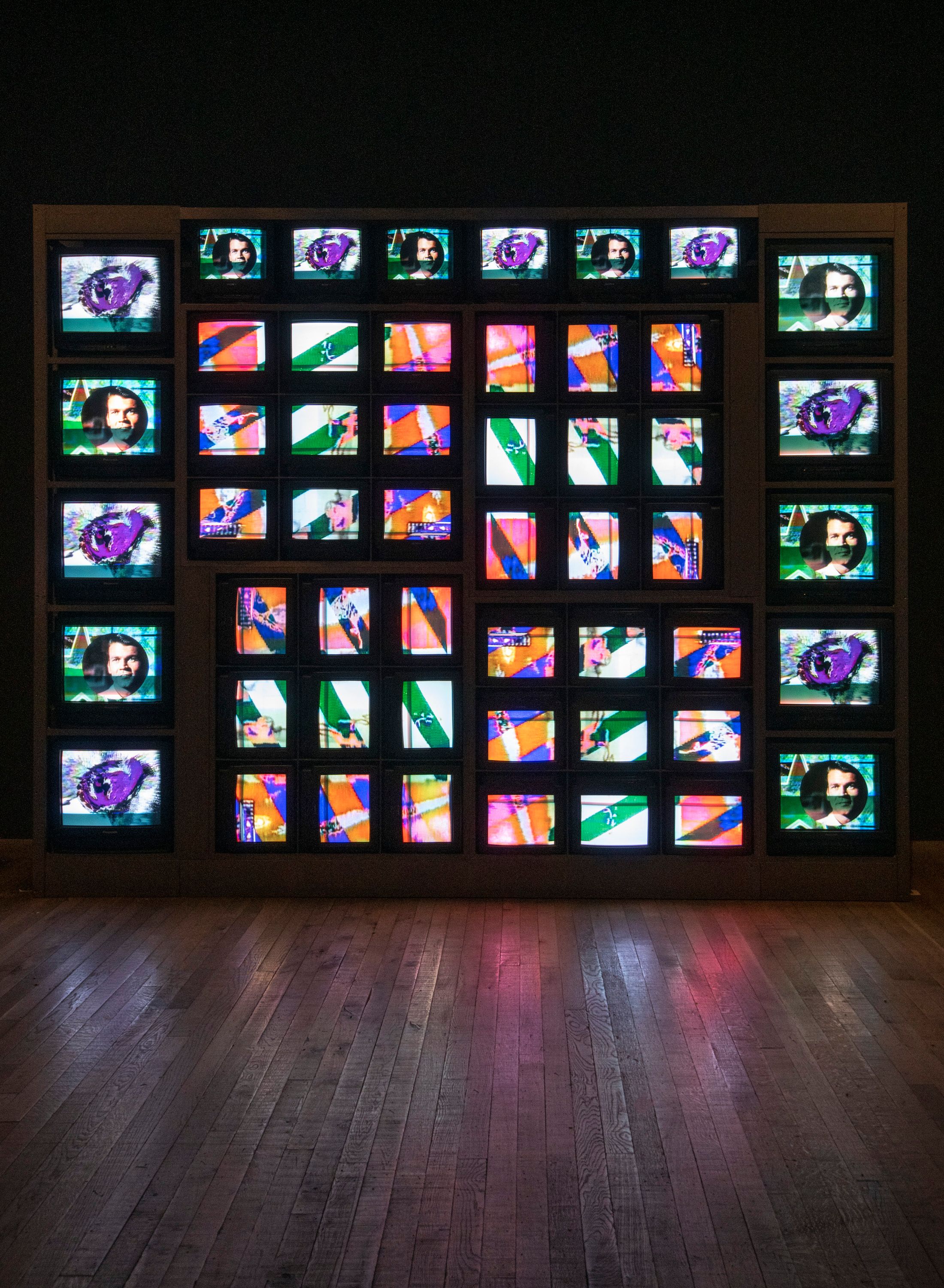 An assortment of televisions and computer screens stacked together, each reflecting different sections of a human face. Th...