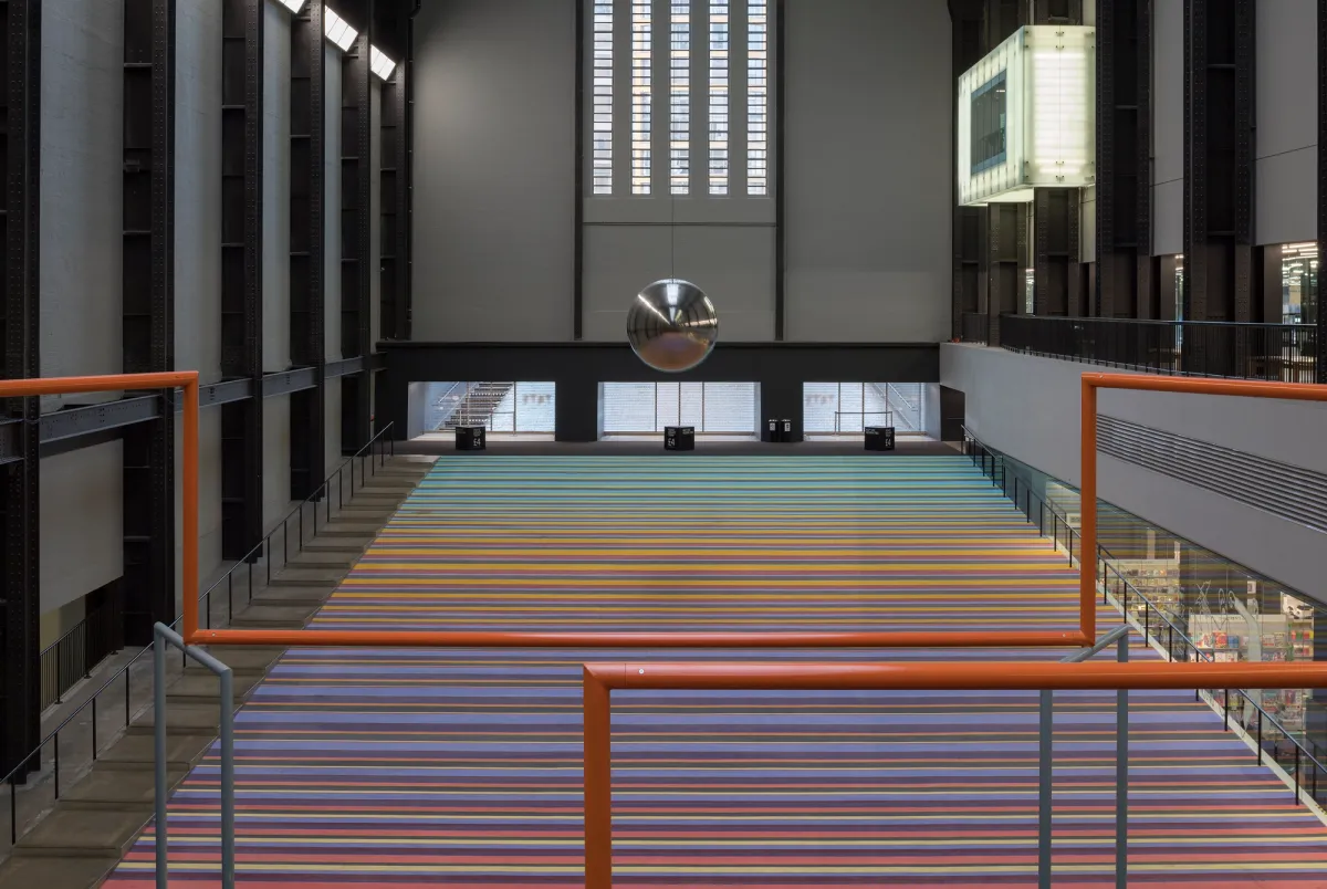 An expansive indoor space with towering windows illuminating orange pipes that traverse the room. Steps lead upward in the distance.