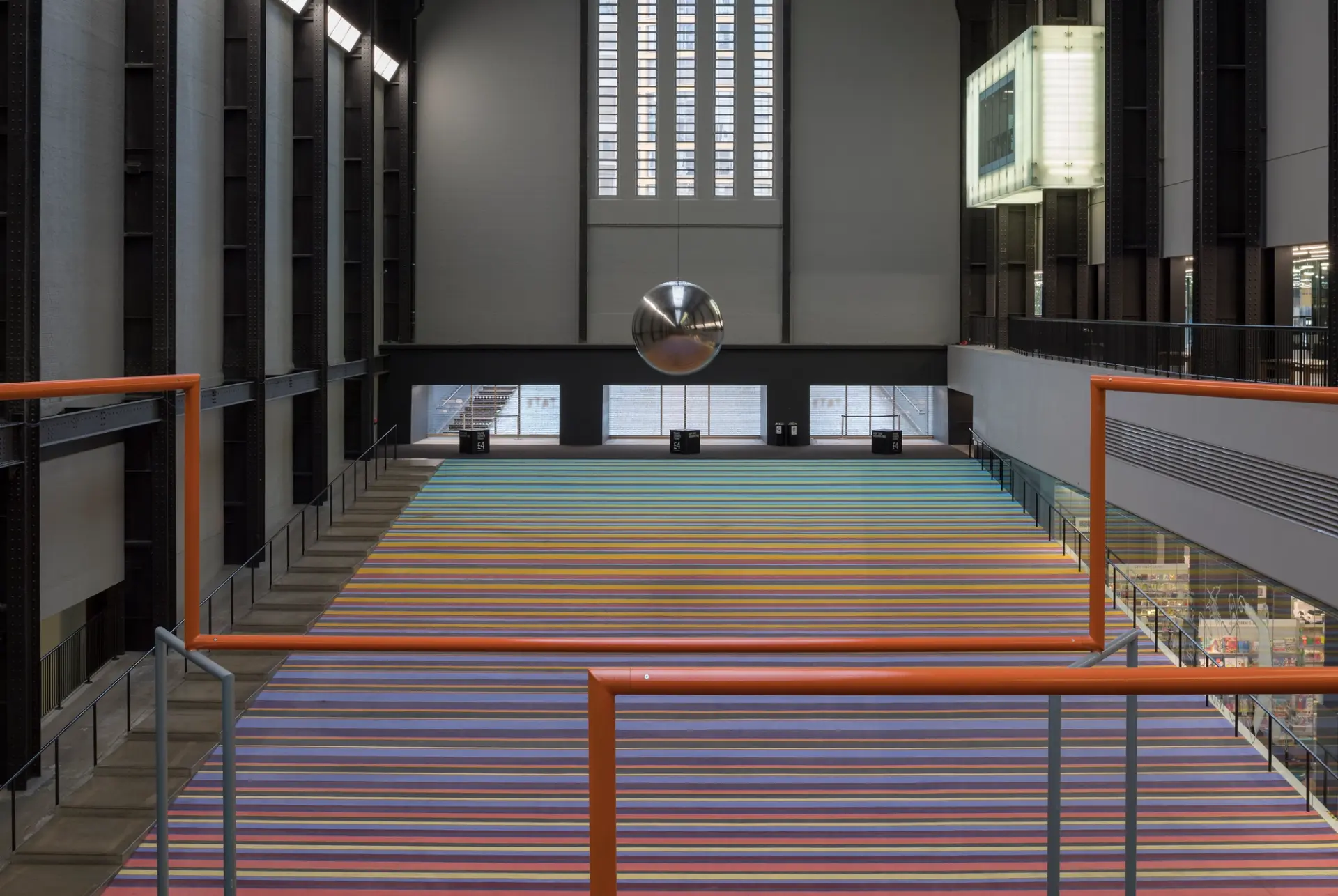An expansive indoor space with towering windows illuminating orange pipes that traverse the room. Steps lead upward in the...
