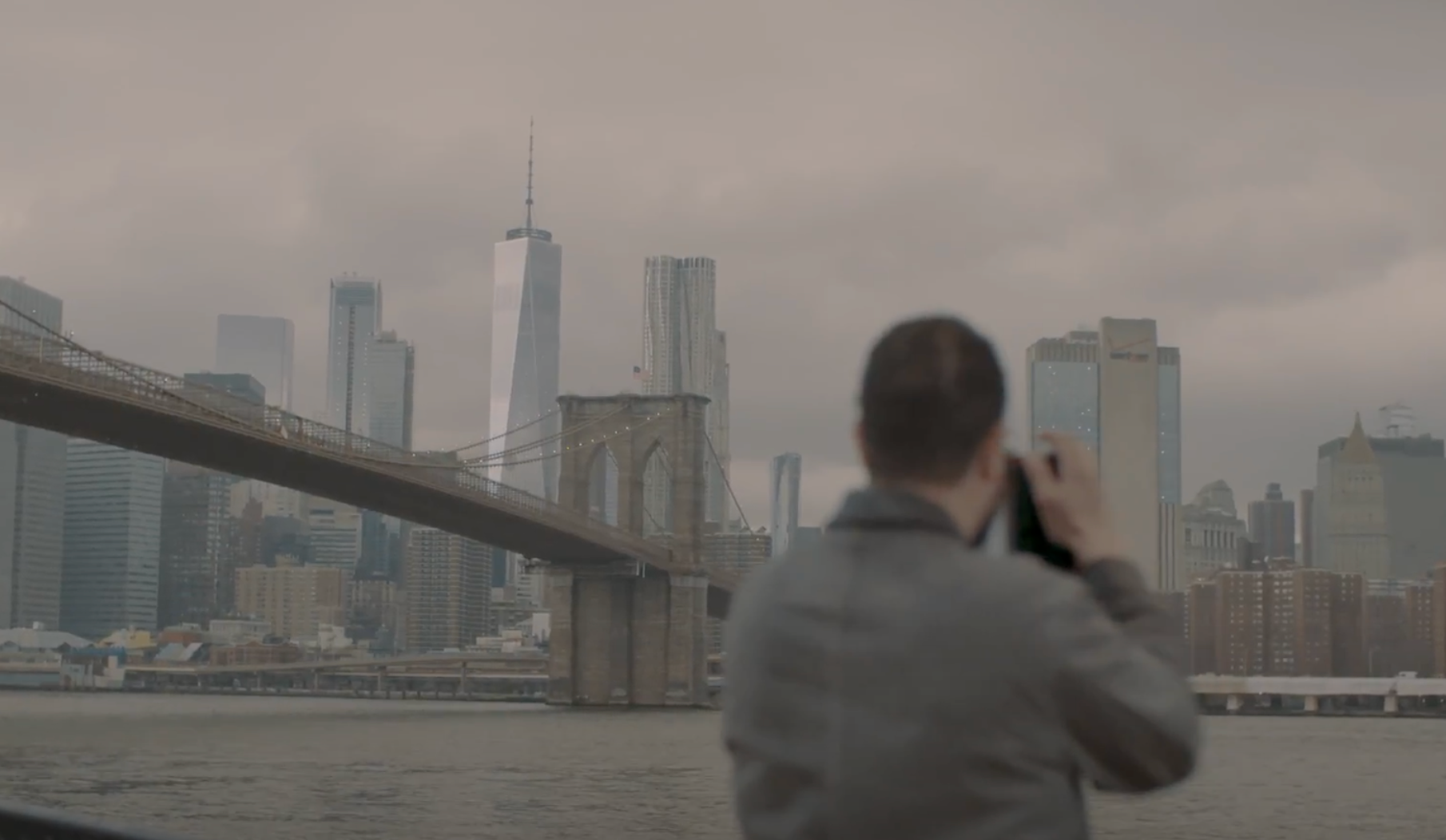 A person stands at a distance capturing a photograph of the Brooklyn Bridge. The weather is cloudy, casting a grey hue ove...