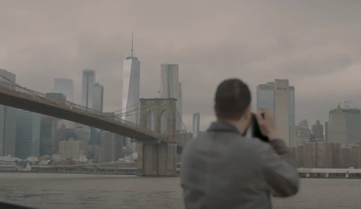 A person stands at a distance capturing a photograph of the Brooklyn Bridge. The weather is cloudy, casting a grey hue over the scene. The iconic bridge cables and towers stand tall in the backdrop.