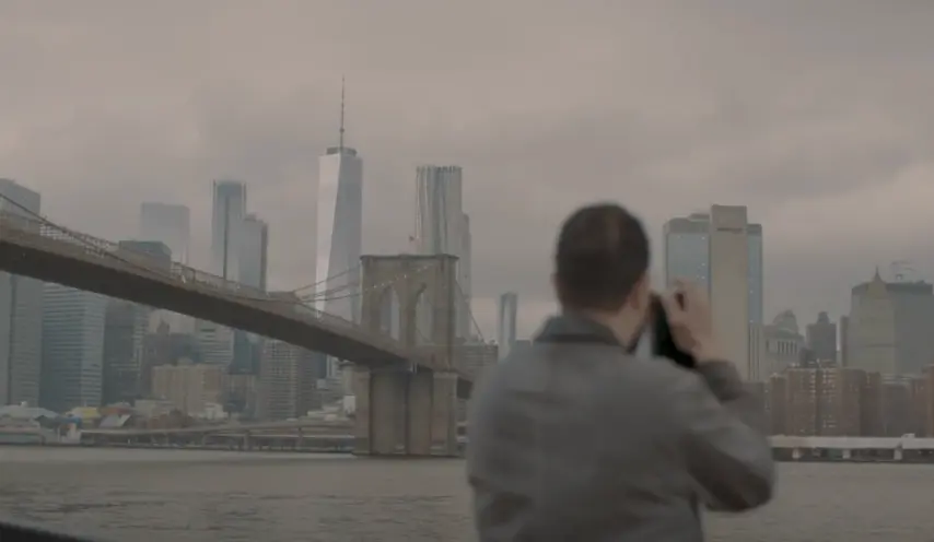 A person stands at a distance capturing a photograph of the Brooklyn Bridge. The weather is cloudy, casting a grey hue ove...
