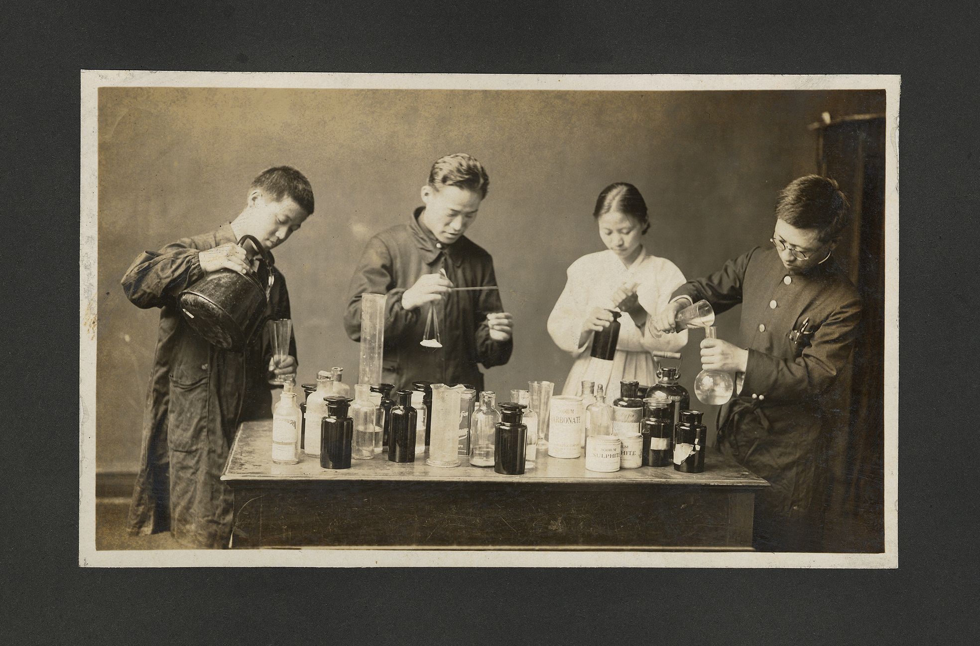 Four chemists, engrossed in experimentation, are gathered around a table laden with multiple glass bottles. The scene is c...