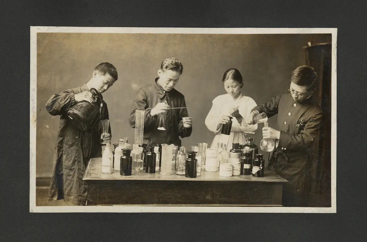 Four chemists, engrossed in experimentation, are gathered around a table laden with multiple glass bottles. The scene is captured in an old sepia-toned photo taken in a 1931 YMCA chemistry lab.
