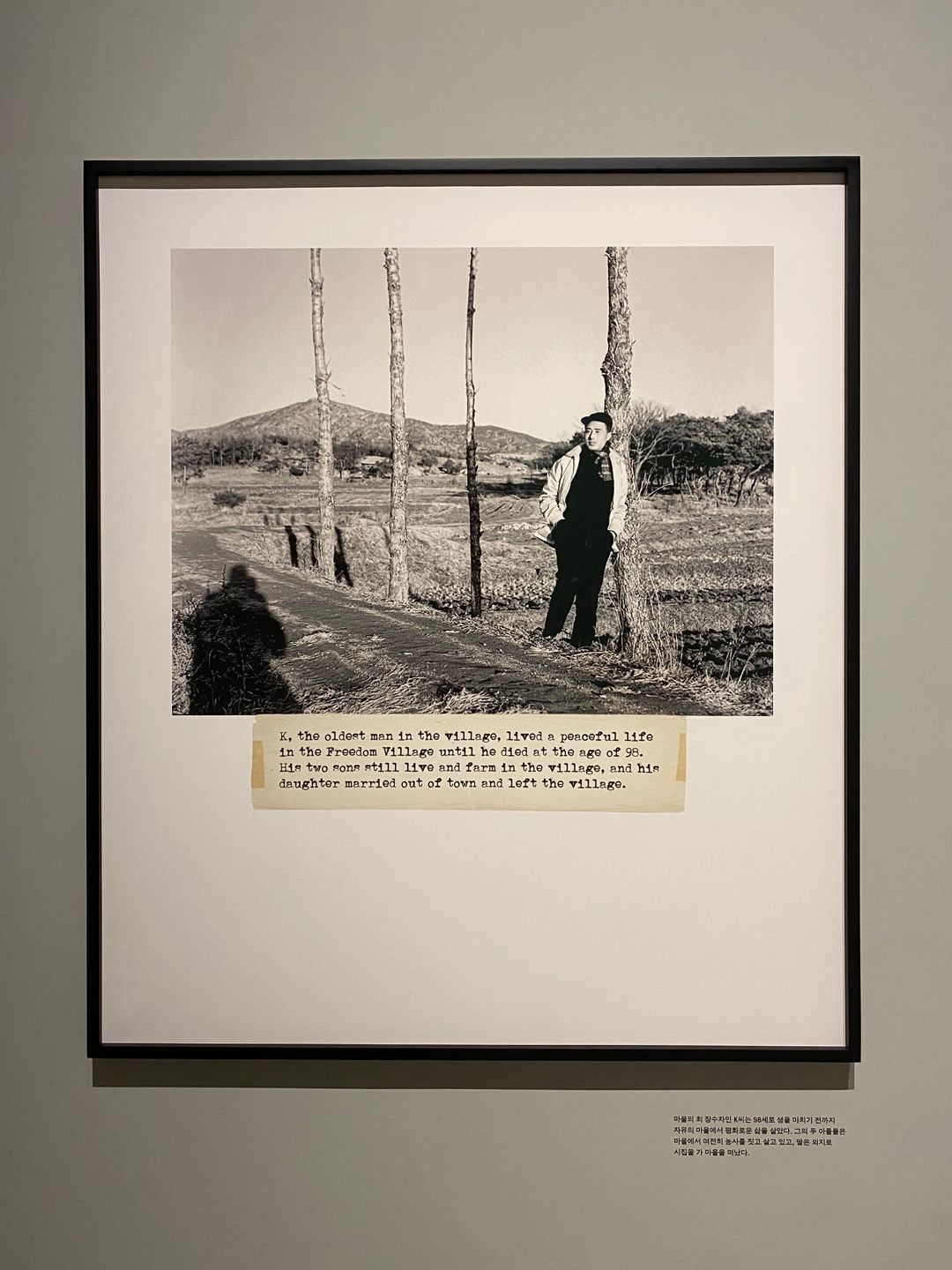 A framed digital print of a vintage photograph featuring an individual leaning on a tree in an open field. Typed caption i...
