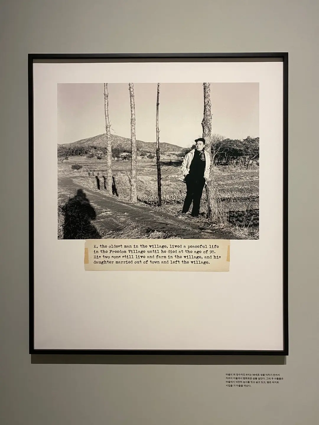 A framed digital print of a vintage photograph featuring an individual leaning on a tree in an open field. Typed caption is just below the image.