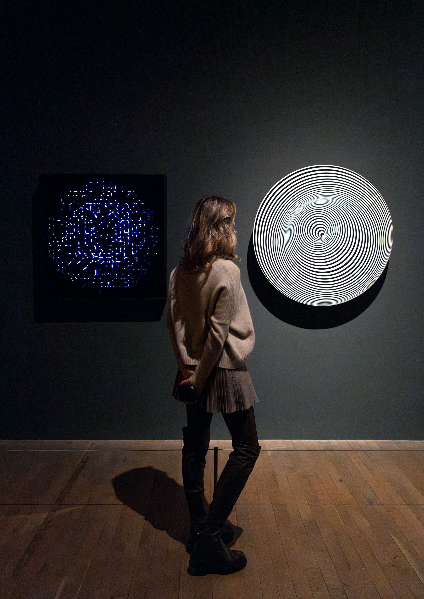 A woman stands in a dimly lit gallery viewing two abstract wall pieces—one a glowing blue grid of dots and lines on a blac...