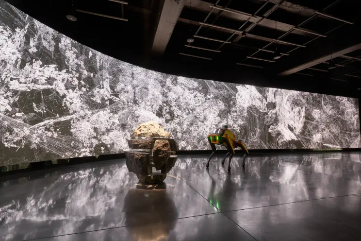 A yellow robotic dog facing a panoramic video display.