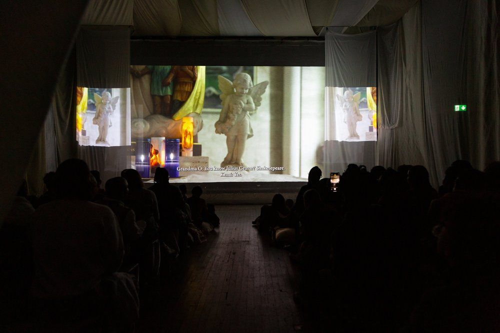 Viewers in a dimly lit room watching projected images, immersed in a cultural or artistic presentation.