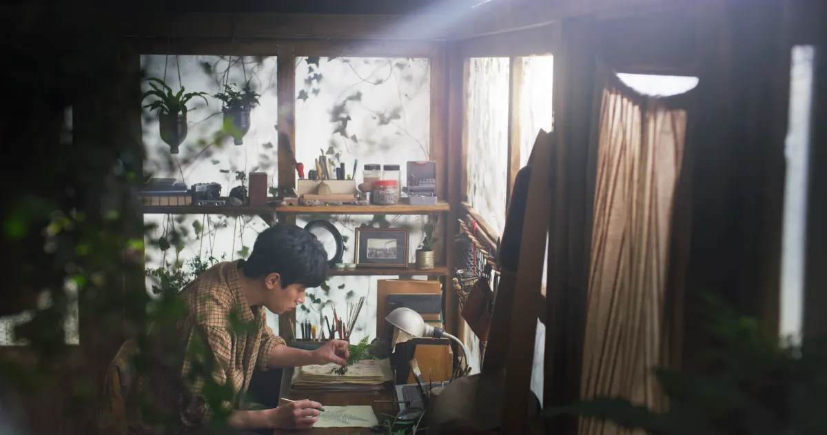 An individual is engrossed in a drawing task at a desk, surrounded by verdant houseplants, in a film still from NEWS FROM NOWHERE: FREEDOM VILLAGE.