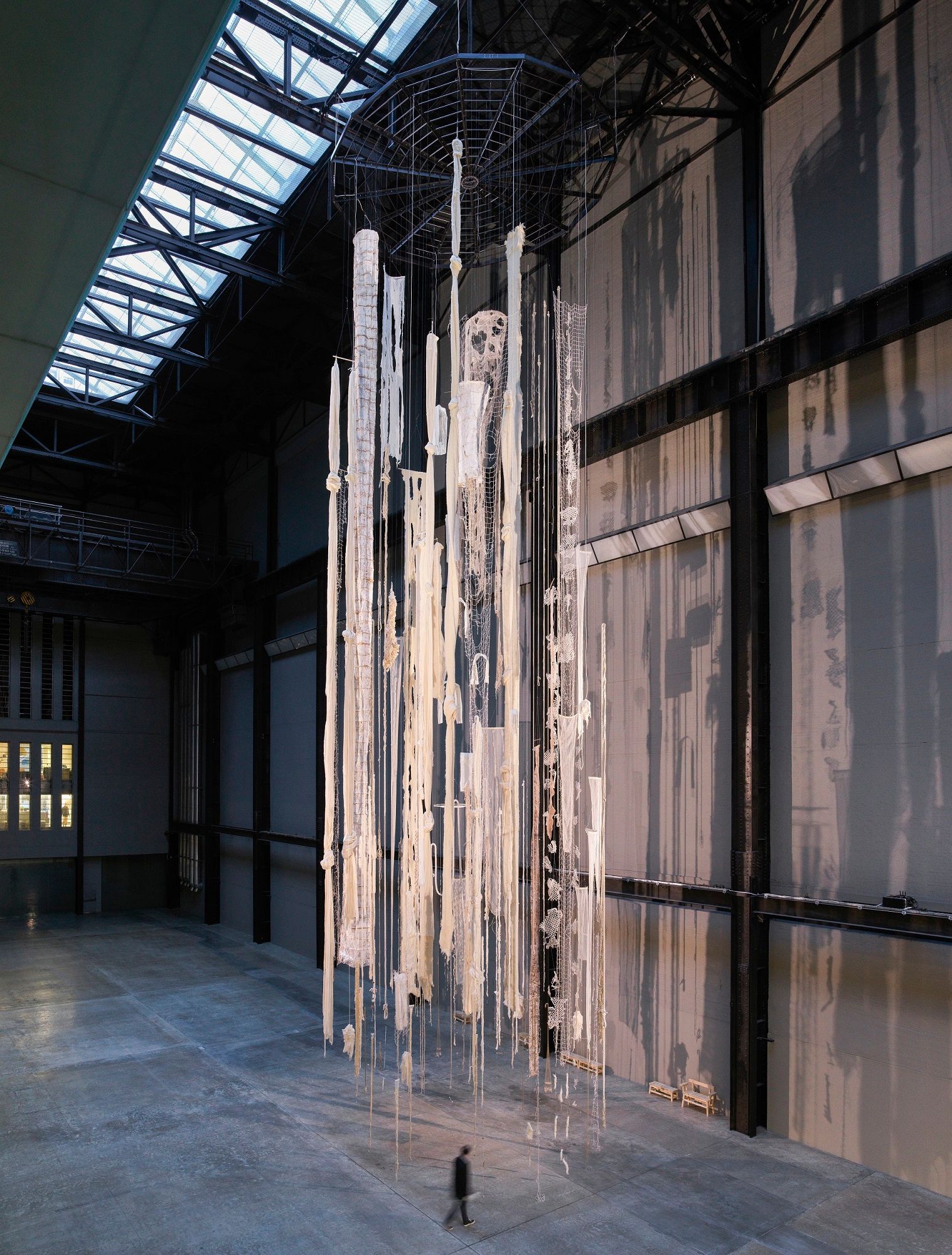 A high-ceiling gallery hall with white ripped curtains, fishnets, and rugs suspended from above. A person walks below the ...