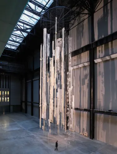 A high-ceiling gallery hall with white ripped curtains, fishnets, and rugs suspended from above. A person walks below the ...