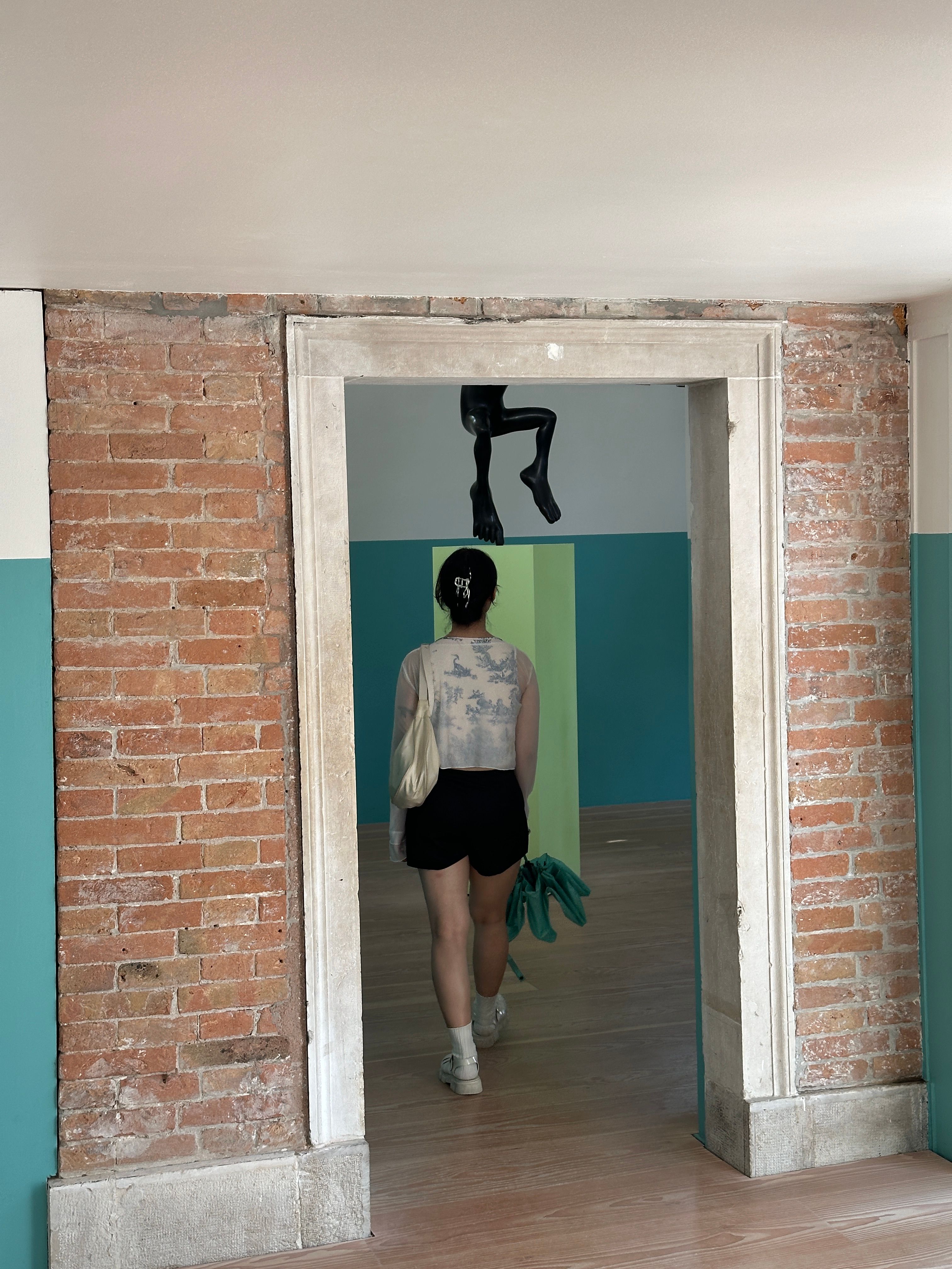 A person walks through a doorway framed by exposed brick and teal-painted walls. Above the doorway, a black sculptural leg is suspended.