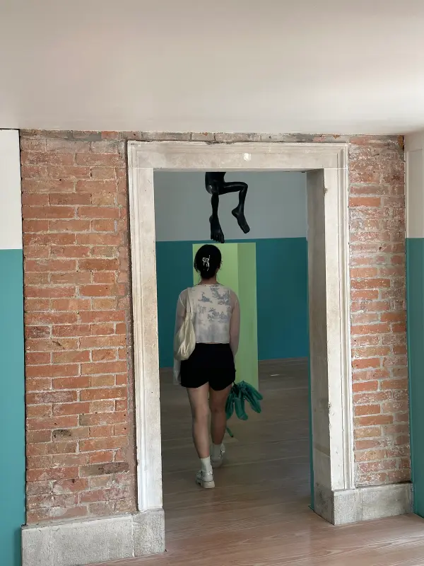 A person walks through a doorway framed by exposed brick and teal-painted walls. Above the doorway, a black sculptural leg is suspended.