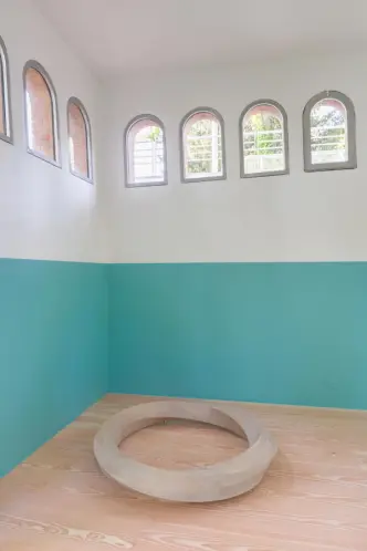 A room with white walls divided by a teal-colored band, arched windows showing the exterior and a large circular wooden ob...