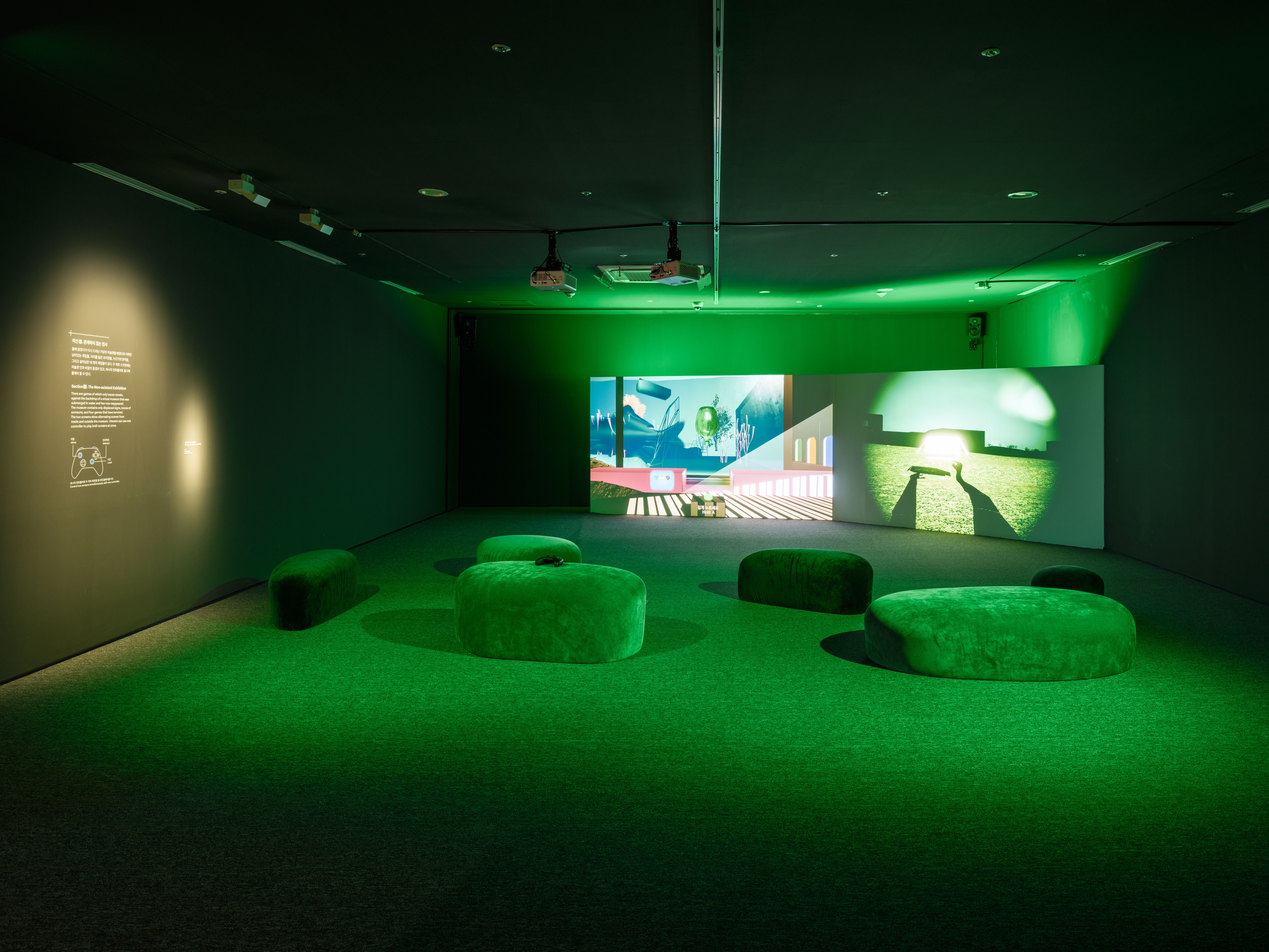 A darkened gallery bathed in green light features a large dual-channel video projection showing a surreal digital landscap...