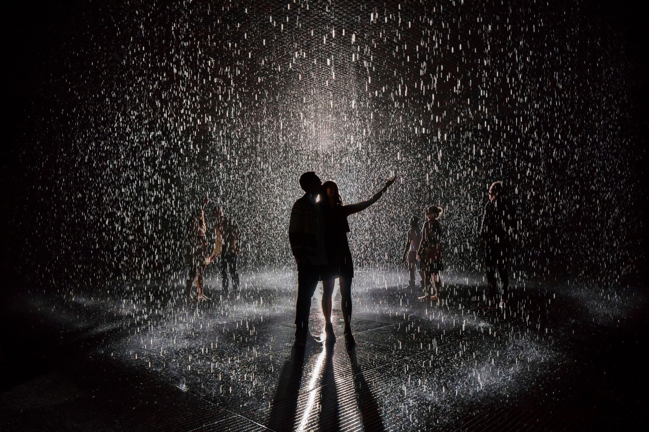 Two silhouettes embracing under a spotlight in a room of heavy rain, surrounded by other figures.