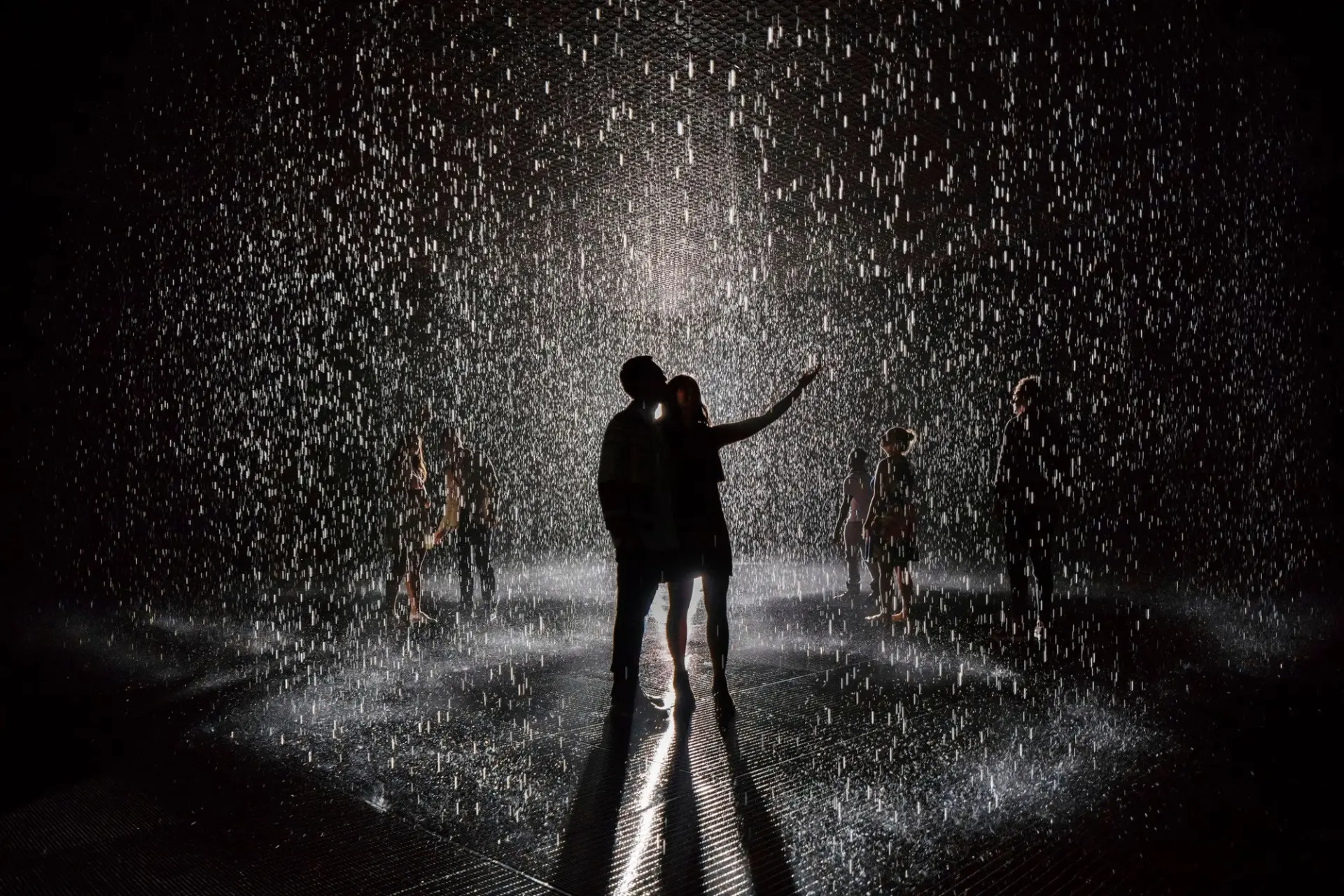 Two silhouettes embracing under a spotlight in a room of heavy rain, surrounded by other figures.