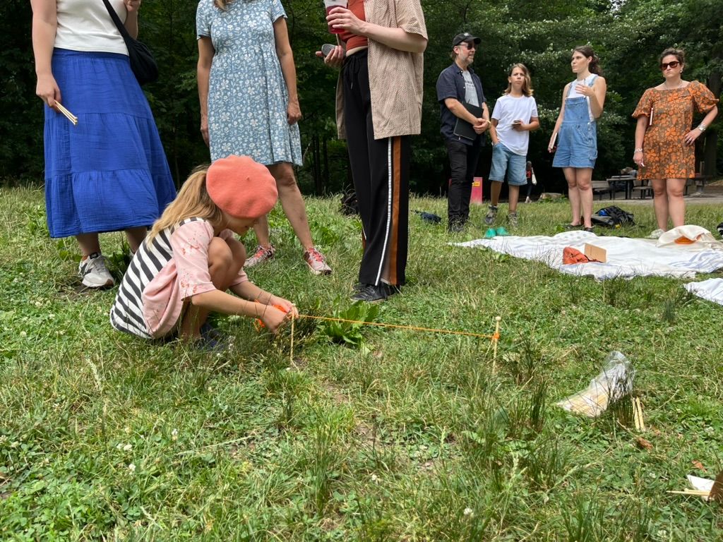 Child crouched on the grass, attaching orange yarn to wooden pegs, with a group of adults nearby.