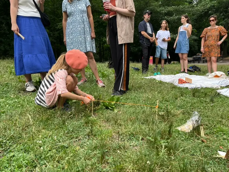 Child crouched on the grass, attaching orange yarn to wooden pegs, with a group of adults nearby.