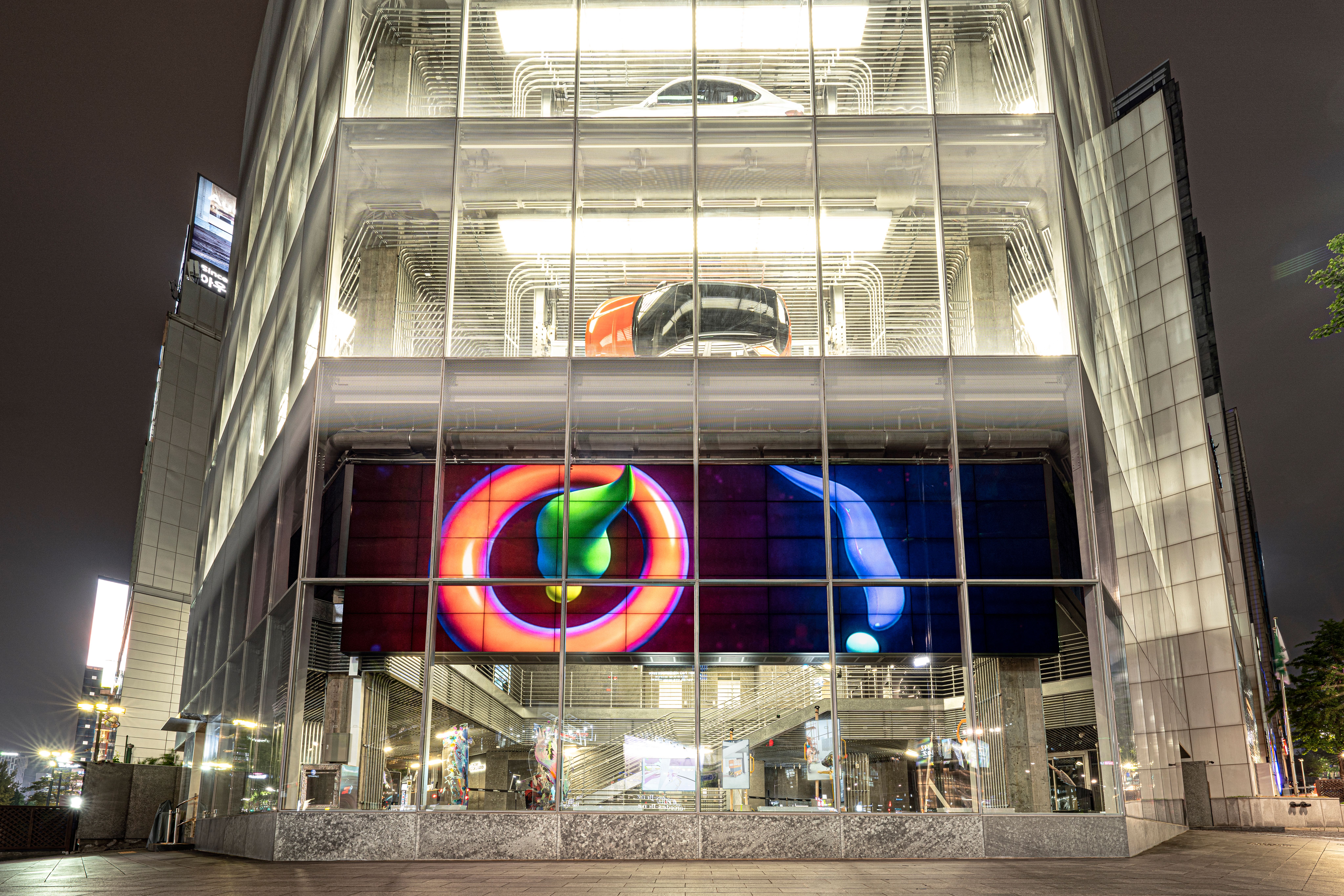 A glass-walled building houses a car exhibit. Screens display various shapes in vibrant colors, adding a dynamic element t...