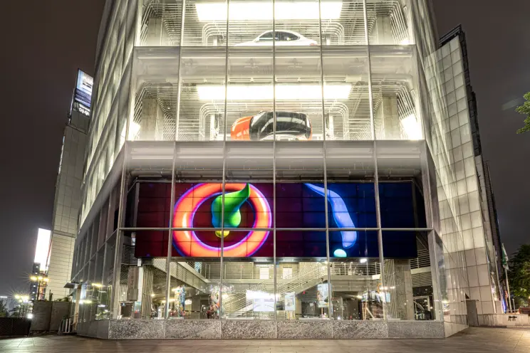 A glass-walled building houses a car exhibit. Screens display various shapes in vibrant colors, adding a dynamic element t...