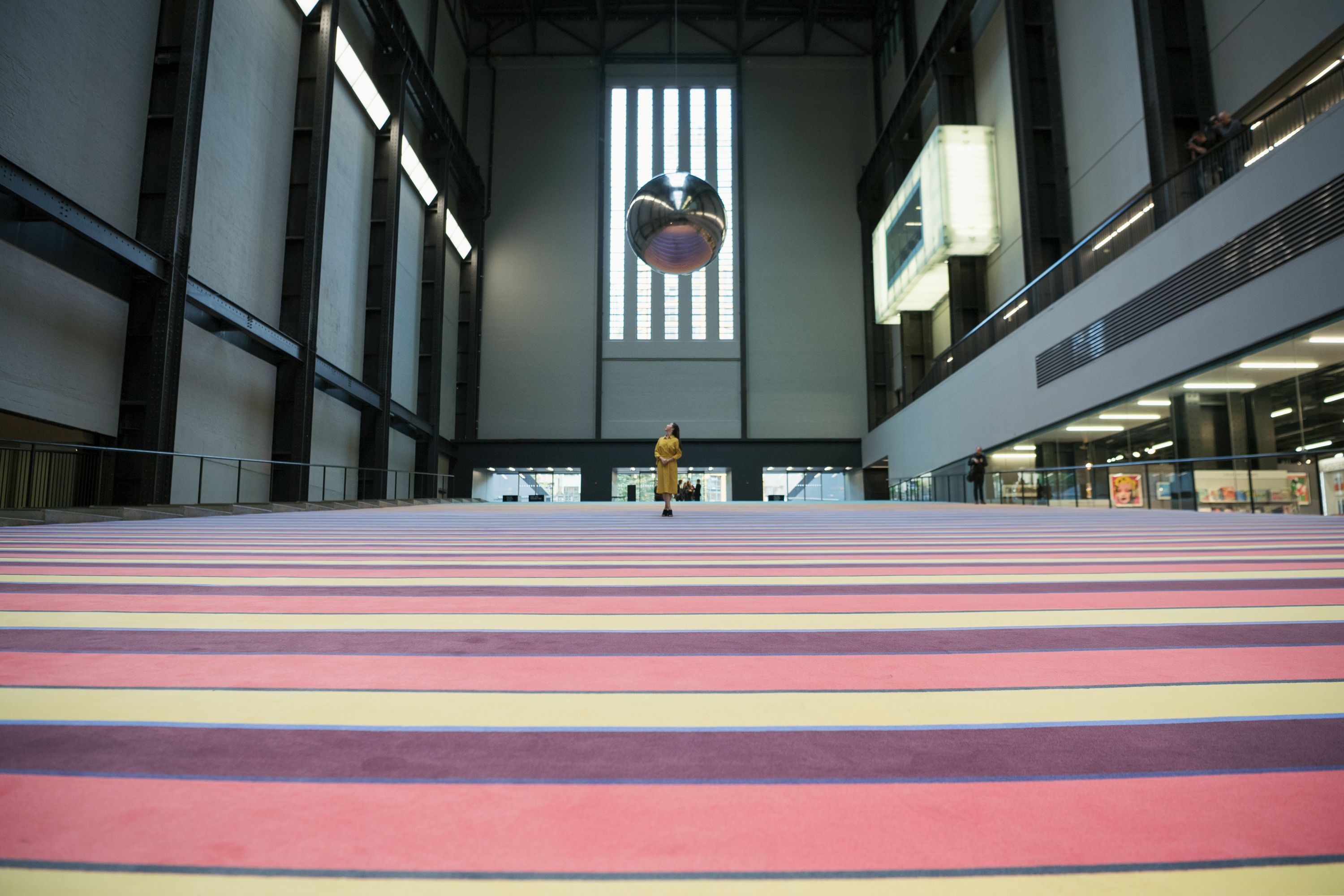 A person in a yellow dress stands on pink stairs and looks up at a metallic ball hanging from the high ceiling.