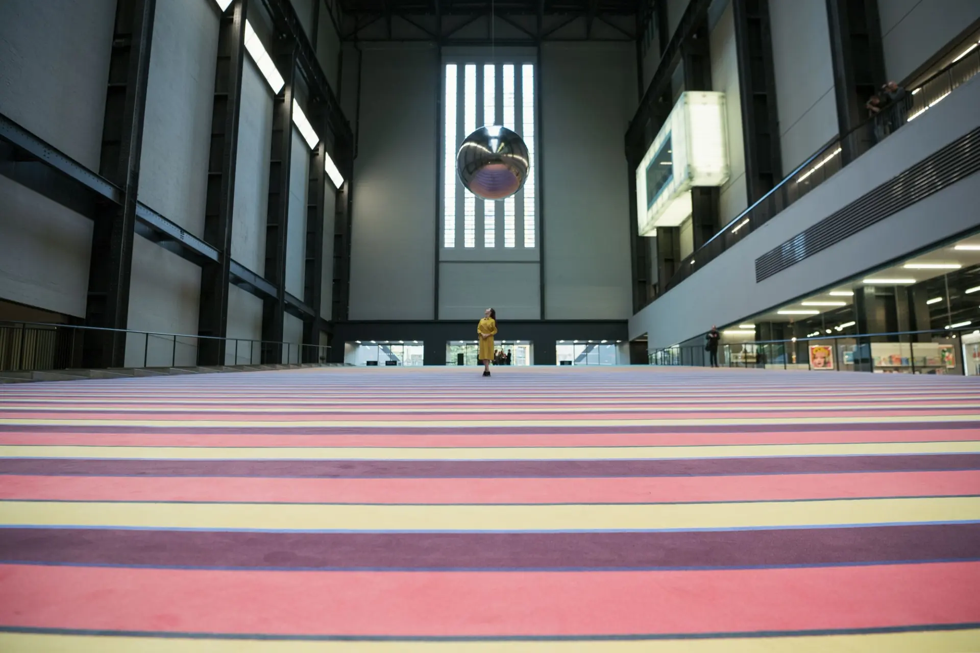 A person in a yellow dress stands on pink stairs and looks up at a metallic ball hanging from the high ceiling.