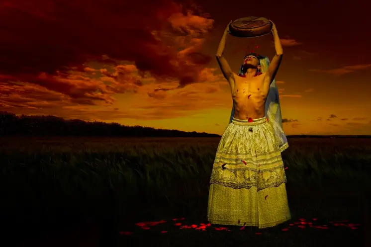 A person in vibrant traditional costume outside at twilight, raising a circular object high above their head.