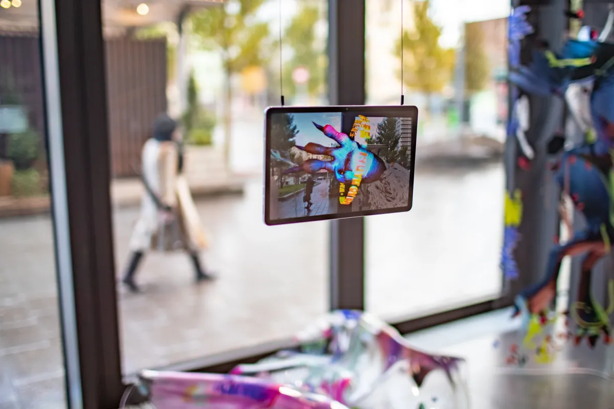 A tablet displaying art is placed near a window, with other art pieces in view. Outside the window, a person is walking on a tree-lined street.